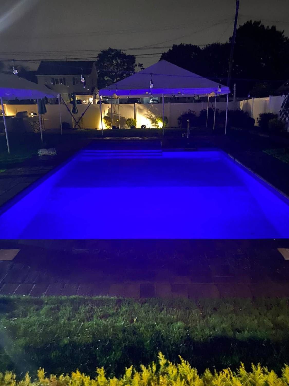 Piscina iluminada de azul de noche, con luces de jardín junto a la cerca